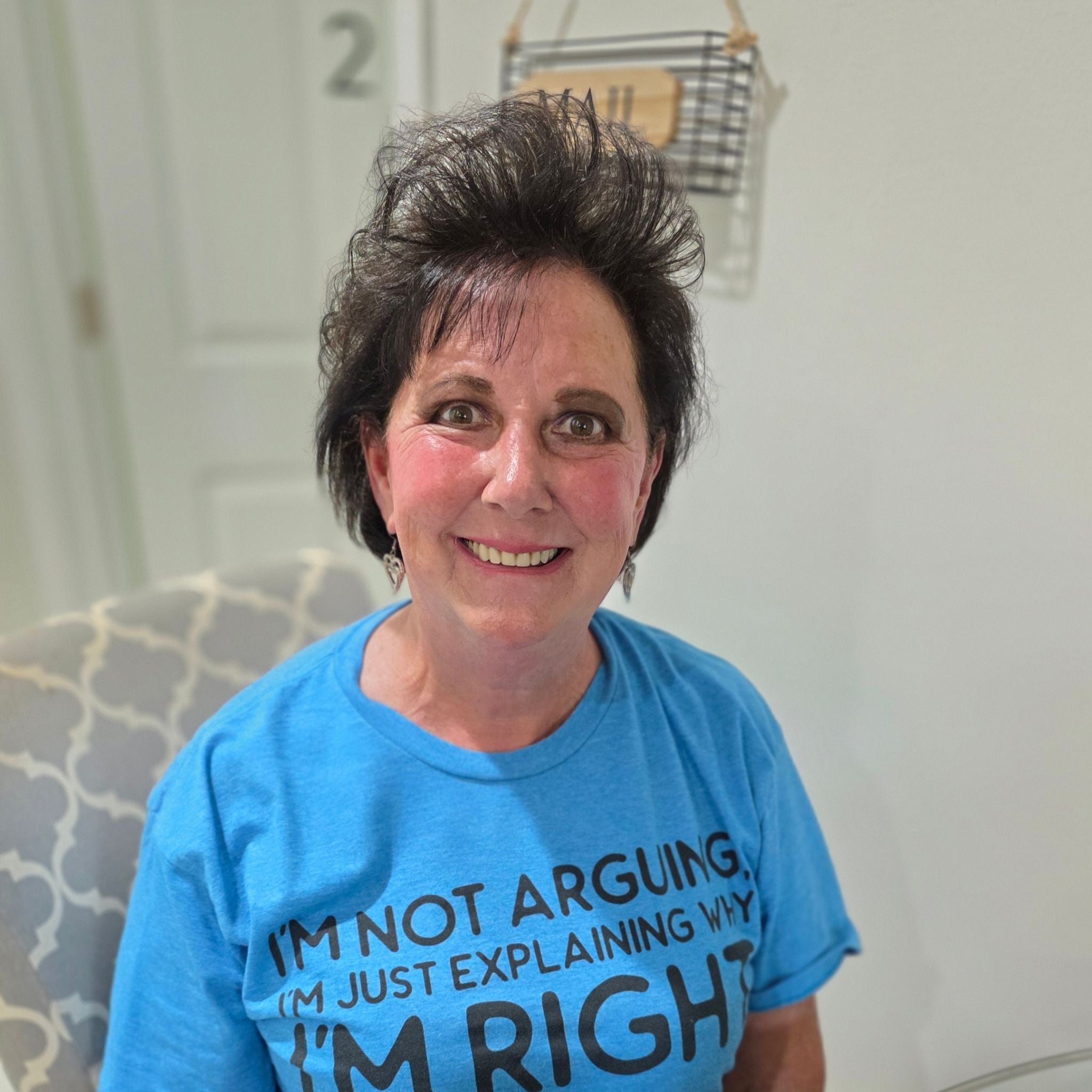 A portrait of an elderly woman named Brenda sitting at a wooden kitchen table and smiling at the camera.